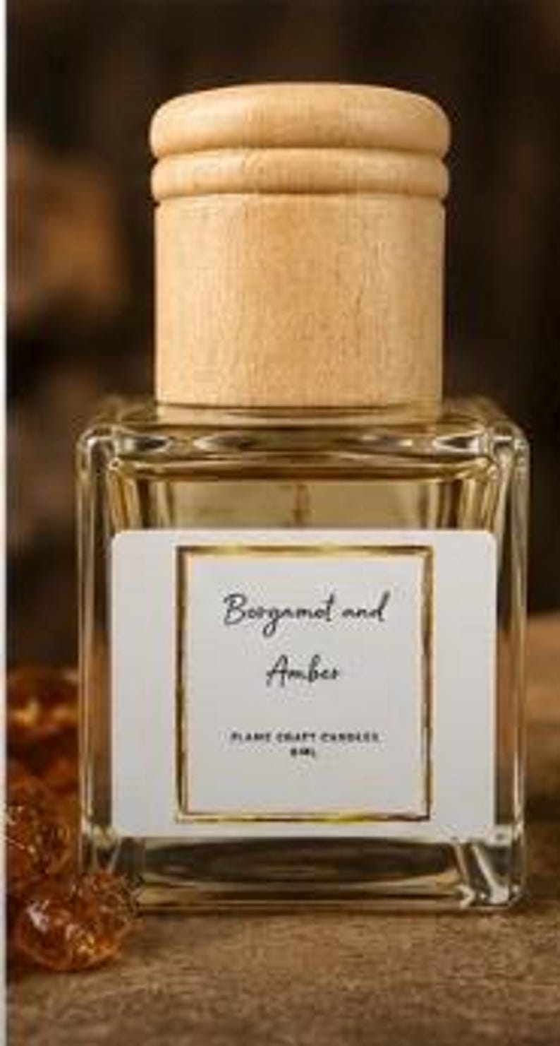 May include: A square glass bottle filled with a light amber liquid, topped with a cylindrical light wood cap. The white label features a gold border and the text Bergamot and Amber, Flame Craft Candles, 60ml.