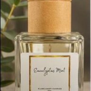 May include: A clear glass square bottle with a light wood cylindrical cap. The white label features a gold border and the text Eucalyptus Mint, Flame Craft Candles, and 8FL. The bottle contains a clear liquid and is topped with a wooden lid.