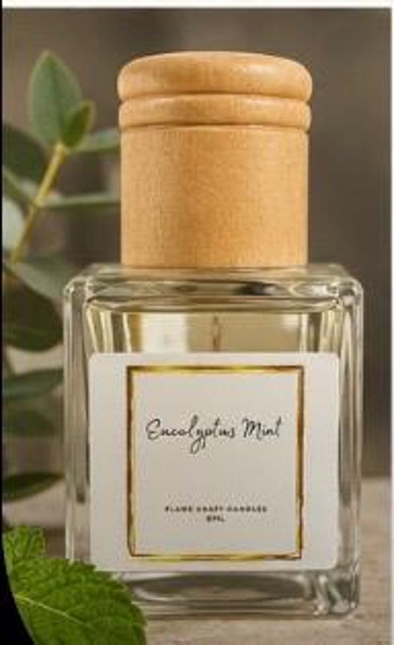 May include: A clear glass square bottle with a light wood cylindrical cap. The white label features a gold border and the text Eucalyptus Mint, Flame Craft Candles, and 8FL. The bottle contains a clear liquid and is topped with a wooden lid.