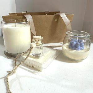 Op de afbeelding: Een verzameling geurkaarsen en accessoires. Inclusief een grote witte kaars in een glas, een kleine blauwe kaars in een pot, een kleine glazen fles en wax melts. Een bruine papieren cadeauzak met strikhandvatten staat op de achtergrond.