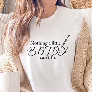 Op de afbeelding: Wit T-shirt met een zwarte afbeelding met de tekst "Nothing a little BOTOX can't fix" met een injectiespuit-illustratie.