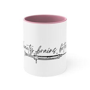 May include: White ceramic mug with a pink rim. The mug features a black and white illustration of a syringe with the text "beauty, brains, botox" written above it.
