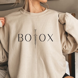 May include: A beige sweatshirt with the word "BOTOX" printed in black with a stylized syringe graphic.