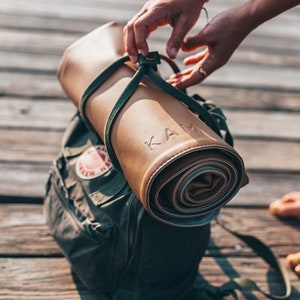 May include: A rolled-up brown leather yoga mat with the word "KAM" embossed on it. The mat is secured with a green strap and is being placed into a green backpack.