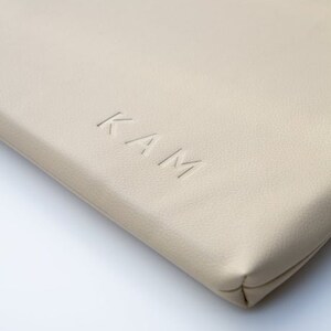 K&ouml;nnte beinhalten: Nahaufnahme einer beige Ledertasche mit dem gepr&auml;gten Wort "KAM" auf der Vorderseite.
