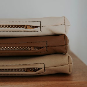 K&ouml;nnte beinhalten: Drei Rei&szlig;verschlusstaschen &uuml;bereinander gestapelt. Die oberste Tasche ist cremefarben, die mittlere Tasche ist braun und die unterste Tasche ist in einem helleren Braunton gehalten. Alle drei Taschen haben goldfarbene Rei&szlig;verschl&uuml;sse.
