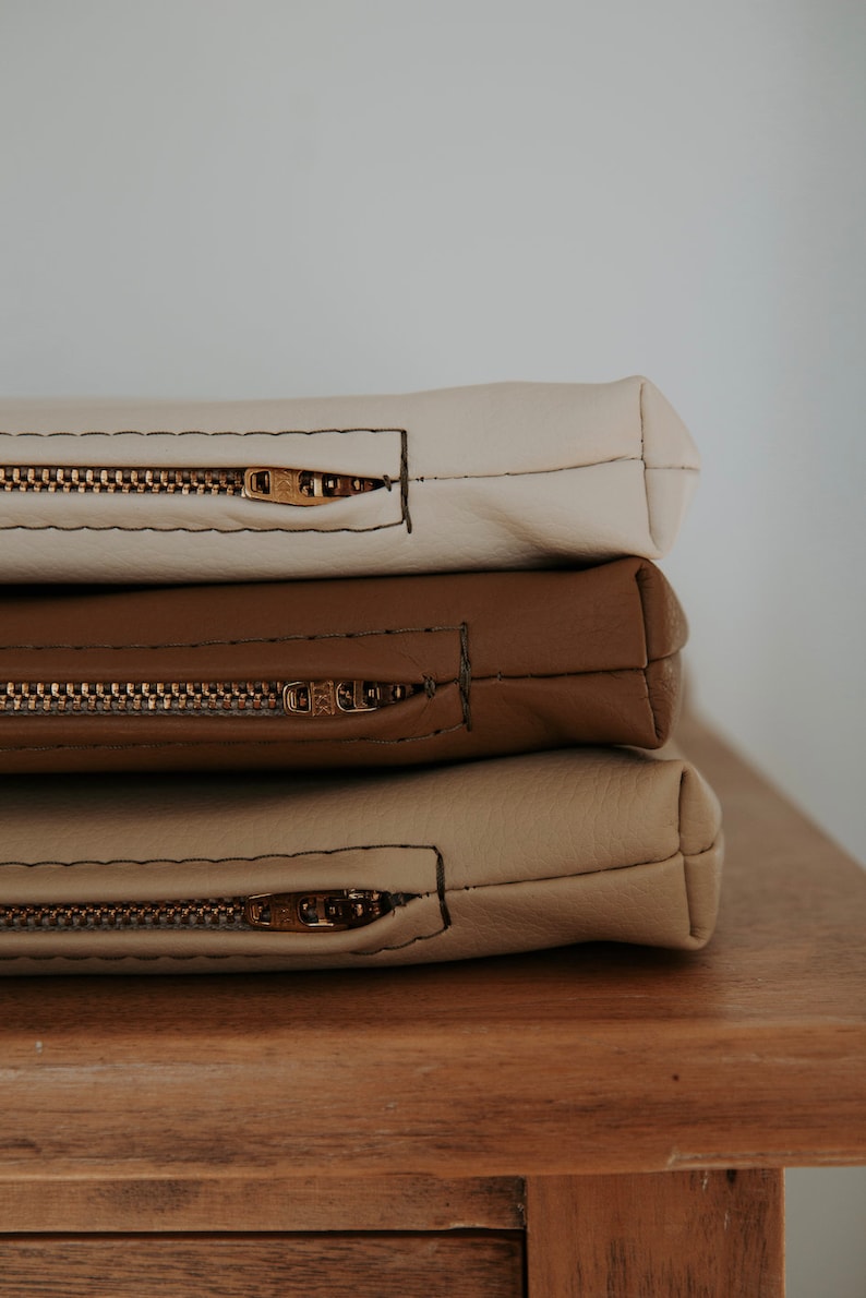 K&ouml;nnte beinhalten: Drei Rei&szlig;verschlusstaschen &uuml;bereinander gestapelt. Die oberste Tasche ist cremefarben, die mittlere Tasche ist braun und die unterste Tasche ist in einem helleren Braunton gehalten. Alle drei Taschen haben goldfarbene Rei&szlig;verschl&uuml;sse.