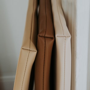 May include: Three leather pouches in different shades of brown, beige, and cream, stacked on top of each other. The pouches are lined up against a white wall.