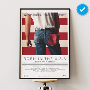 Puede incluir: Un póster para el álbum "Born in the U.S.A." de Bruce Springsteen. El póster presenta una foto de Springsteen de pie frente a una bandera estadounidense, con jeans azules y una camiseta blanca. El título del álbum está impreso en letras grandes y negritas en la parte inferior del póster.