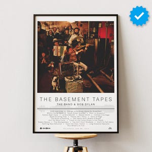 May include: A vintage poster for "The Basement Tapes" by The Band & Bob Dylan. The poster features a black and white photo of the band in a basement setting. The album title and artist names are printed in white text on a black background. The tracklist is printed below the photo.