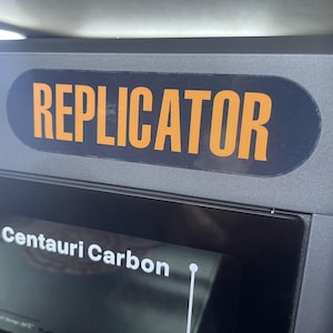 May include: A dark gray machine with a black and orange label that reads "REPLICATOR." Below the label is a screen displaying "Centauri Carbon."