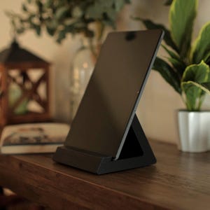 Könnte beinhalten: Ein schwarzer Tablet-Ständer mit dreieckigem Fuß, abgewinkelt zur Aufnahme eines Tablets. Der Ständer steht auf einer dunklen Holzoberfläche, mit einer kleinen weißen Vogelfigur und einer Topfpflanze im Hintergrund.