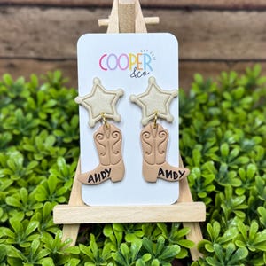 May include: A pair of dangle earrings featuring a cream-colored star stud attached to a tan cowboy boot charm. The boot has intricate embossed detailing and the name ANDY written in black on the side. The earrings are displayed on a white card.
