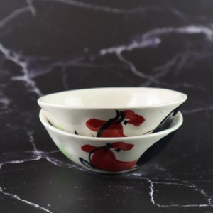 Puede incluir: Dos cuencos de cerámica blanca con diseños florales rojos y negros. Los cuencos están apilados uno encima del otro.