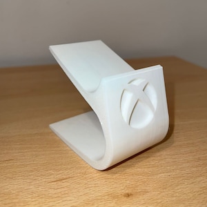 May include: White plastic stand for a video game console. The stand has a cutout of the Xbox logo.