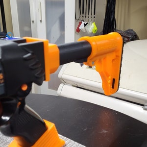 May include: A black and orange toy gun with the text "BUG-A-S THE ORIGINAL V2" printed on the side. The gun has a black barrel and an orange stock.
