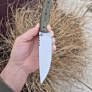 May include: A stainless steel knife with a serrated edge and a green handle. The knife has a straight blade and a small emblem near the handle. The handle is textured and has several black rivets. The knife is held in a hand.