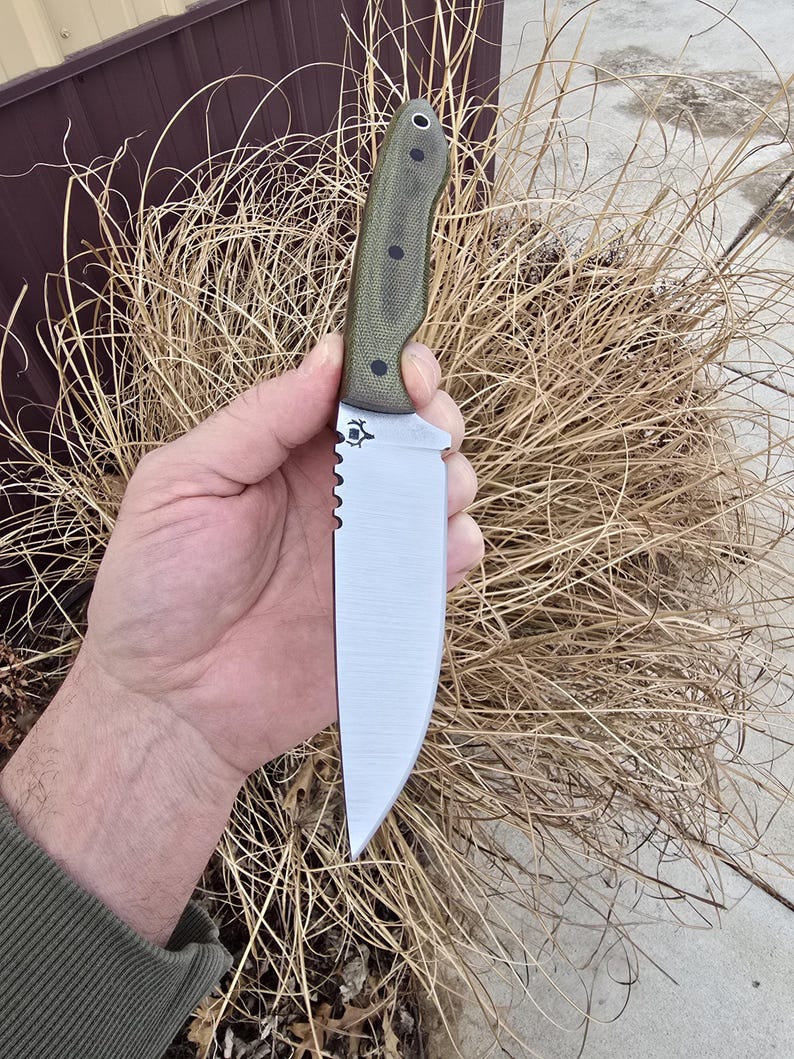 May include: A stainless steel knife with a serrated edge and a green handle. The knife has a straight blade and a small emblem near the handle. The handle is textured and has several black rivets. The knife is held in a hand.