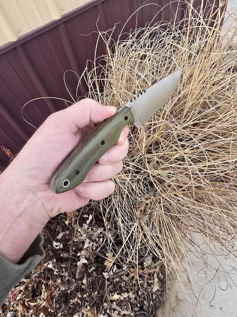 May include: A tactical knife with a silver blade and a textured, olive green handle. The blade has a serrated section near the handle. The knife is held in a hand against a backdrop of dry, brown grass.