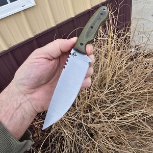 May include: A large, silver-bladed knife with a serrated edge and a green handle. The knife has a pointed tip and a decorative emblem near the handle. The handle is textured with black dots. The knife is held in a hand.