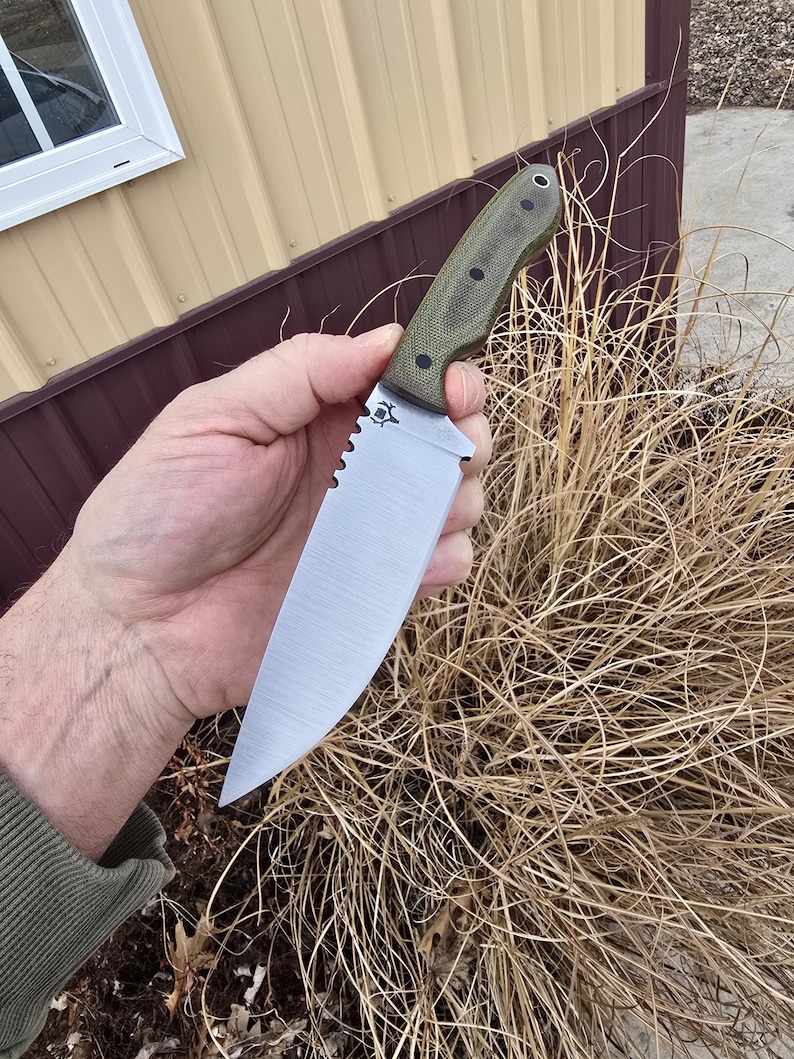 May include: A large, silver-bladed knife with a serrated edge and a green handle. The knife has a pointed tip and a decorative emblem near the handle. The handle is textured with black dots. The knife is held in a hand.