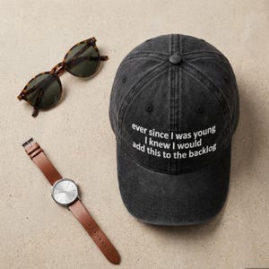 May include: A flat lay featuring a black baseball cap with white text, tortoiseshell sunglasses, and a silver watch with a brown leather strap. The cap reads "ever since I was young I knew I would add this to the backlog."