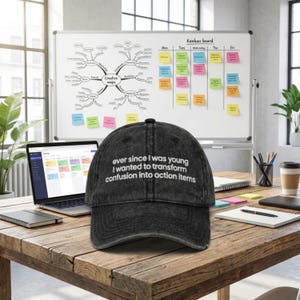 May include: A black denim baseball cap with the text "ever since I was young I wanted to transform confusion into action items" in white. The cap is on a wooden desk with a laptop, whiteboard, and coffee cup.