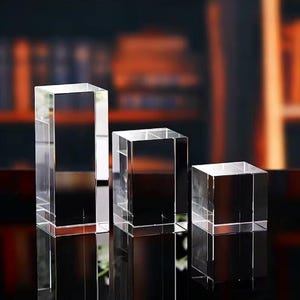 May include: Three clear, rectangular crystal blocks of varying heights. The blocks are arranged on a reflective surface, with the tallest block on the left and the shortest on the right. The background is blurred, with warm tones.