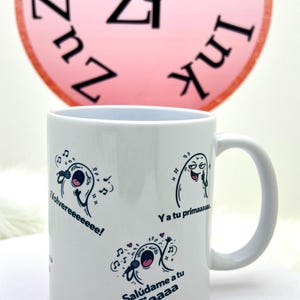 May include: White ceramic mug featuring cartoon ghost-like characters singing into microphones. The mug has the phrases "Volvereeeeeee!", "Ya tu primaaaaa", and "Salúdame a tu Tiaaaa" printed on it. A pink circle with black text is visible in the background.