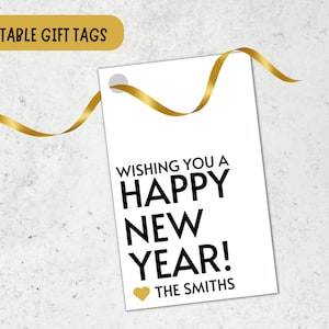 Op de afbeelding: Witte cadeaukaart met zwarte tekst die "Wishing you a Happy New Year!" luidt, met een gouden hart en de tekst "The Smiths" eronder. De kaart is vastgebonden met een gouden lint.