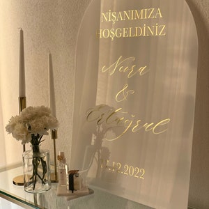 Puede incluir: Un arco de acrílico blanco con letras doradas. El letrero dice "NİŞANIMIZA HOŞGELDİNİZ" y "Nura & Ertuğrul" con la fecha "17.12.2022".