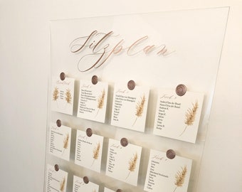May include: A clear acrylic seating chart with a handwritten inscription "Sitzplan" in rose gold script. The chart is divided into sections labeled "Brauttisch", "Tisch 1", "Tisch 2", etc. Each section has a small card with a list of names and a decorative dried grass element.