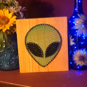 May include: A wooden string art piece featuring an alien head design. The alien's head is green with black eyes, and the piece is mounted on a light-colored wooden board. A blue bottle with painted sunflowers and fairy lights is in the background.