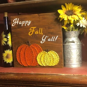 May include: A wooden sign with the text "Happy Fall Y'all!" in white lettering. The sign is decorated with two string art pumpkins, one orange and one yellow. The sign is sitting on a shelf with a bottle of wine and a metal milk jug with sunflowers.
