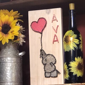 May include: A wooden sign with a string art design of an elephant holding a pink heart balloon. The name "AVA" is spelled out in string art on the right side of the sign.