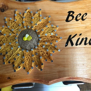 May include: A wooden sign with a yellow string art sunflower and a bee in the center. The sign says "Bee Kind" in black lettering.