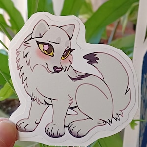 May include: A white wolf sticker with yellow eyes and pink cheeks. The wolf is sitting with its tail curled around its legs.