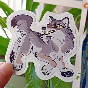 May include: A sticker of a cartoon wolf with a gray and white fur pattern. The wolf is looking to the right with its mouth open, showing its teeth. The wolf has a yellow eye and a blue eye.