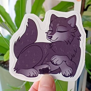 May include: A sticker featuring a cartoon wolf with a purple-grey color scheme. The wolf is lying down with its eyes closed, appearing to be sleeping. The sticker has a white background.