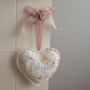 Peut inclure: Ornement en forme de cœur en tissu blanc cassé, brodé du nom "Lily" en cursive. Il est orné de broderies florales et est suspendu à un ruban rose noué en nœud. L'ornement est fixé à une surface blanche.