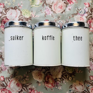 May include: Three vintage white metal canisters with silver lids. The canisters are labeled "suiker", "koffie", and "thee" in black text. The background features a floral pattern.