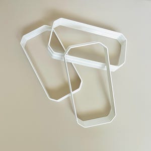 May include: Three white rectangular cookie cutters with beveled edges are arranged on a neutral background. The cutters are made of a solid, opaque material and are designed with a geometric shape. The cutters are likely used for baking or crafting.