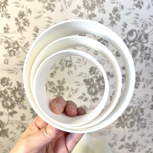 May include: Three white plastic cake rings, nested together, with the largest ring on the bottom and the smallest ring on top.