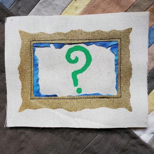 May include: A white canvas with a gold frame and a blue inner frame. A green question mark is painted inside the frame.