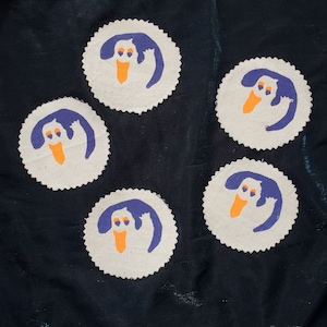 May include: Five white fabric coasters with a purple ghost design. Each ghost has orange eyes and a mouth. The coasters are on a dark blue fabric background.