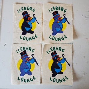 May include: Four square fabric patches with a penguin wearing a top hat and holding a cane. The penguin is on a yellow circle with the text "ICEBERG LOUNGE" in green letters.