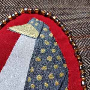 May include: A close-up of a red fabric circle with a black and gold patterned bird design. The circle is surrounded by a beaded trim with brown and silver beads.