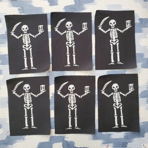 Puede incluir: Seis parches de tela negra con figuras de esqueletos blancos sosteniendo relojes de arena. Cada parche presenta un diseño de calavera y huesos cruzados con el esqueleto sosteniendo un reloj de arena en cada mano.