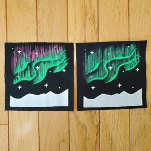 May include: Two square black fabric patches featuring hand-painted designs of the aurora borealis. The left patch displays green and pink light streaks with white stars above a white mountain silhouette. The right patch shows green light streaks.