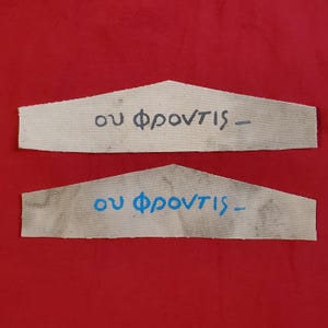 May include: Two beige fabric pieces, each shaped like a house, with the Greek phrase "ou phrontis-" written in blue and black ink. The background is a solid red.